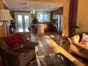 Uma sala de estar ampla com um sofá de couro, uma mesa de centro de vidro e uma poltrona de couro. Uma cozinha com armários de madeira e eletrodomésticos de aço inoxidável é visível ao fundo.