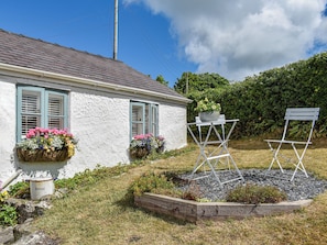A white building with two windows, a flower pot on a stand, and a folding chair.