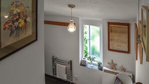 A room with a bed, a chair, a window with plants, a ceiling light, and a framed picture on the wall.