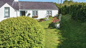 A white house with a grey roof, a chimney, and a small garden with a hedge and a flower pot.