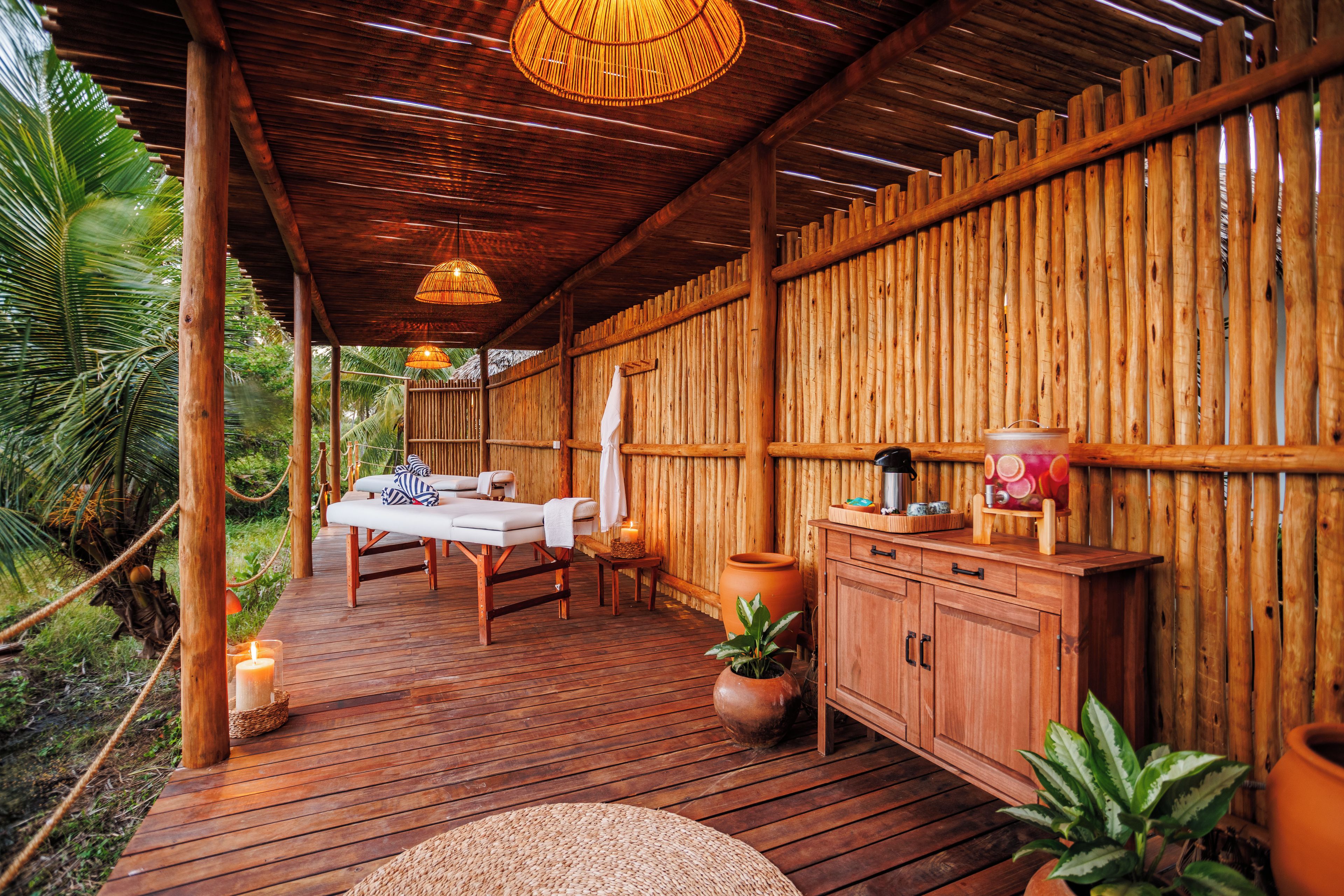 A wooden deck with a massage table and chairs, surrounded by bamboo walls and tropical plants.