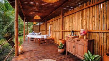 A wooden deck with a massage table and chairs, surrounded by bamboo walls and tropical plants.