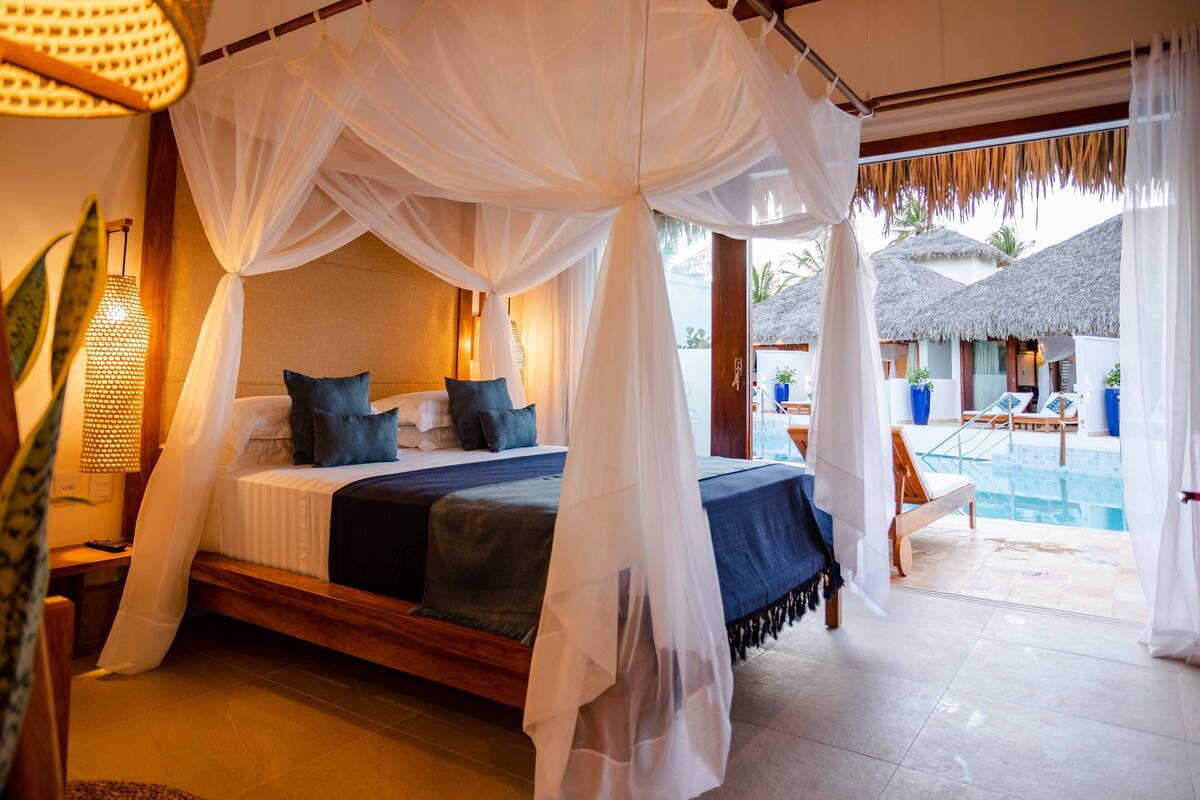 A bedroom with a four-poster bed, a view of the pool, and a thatched-roof structure outside.