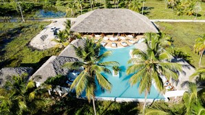 Eine Vogelperspektive zeigt einen luxuriösen Urlaubsort mit einem großen Pool, umgeben von Palmen und strohgedeckten Gebäuden.