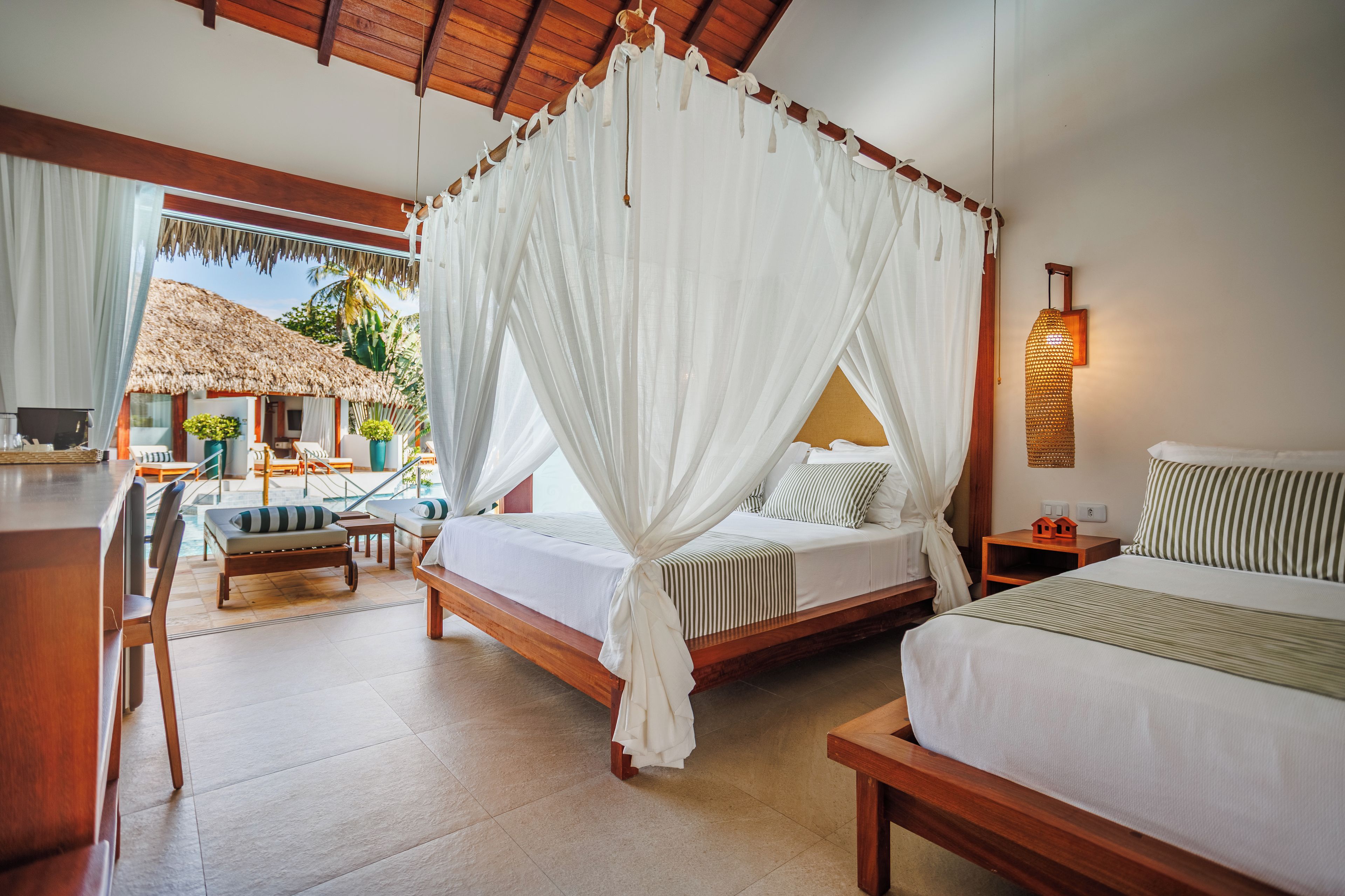 Quarto com cama de dossel, cama extra, mesa pequena, cadeira e vista para a área da piscina.