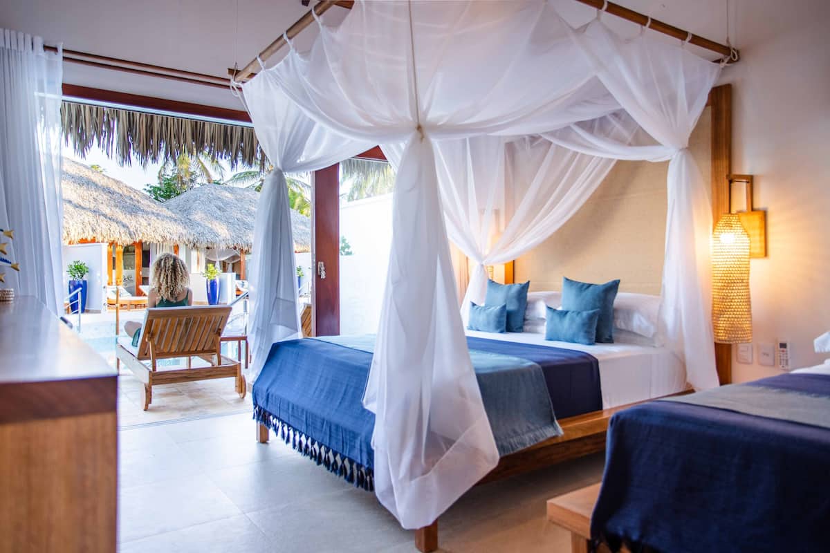 A bedroom with a canopy bed, a separate bed, and a view of a thatched-roof structure outside.