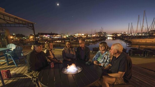 A group of people seated around a round table with a fire pit in the center, overlooking a marina with boats and a cityscape in the background. - Reef Point Marina - Vessel: "Freckled Fin" - East Dock 1, Slip 19 (Racine)