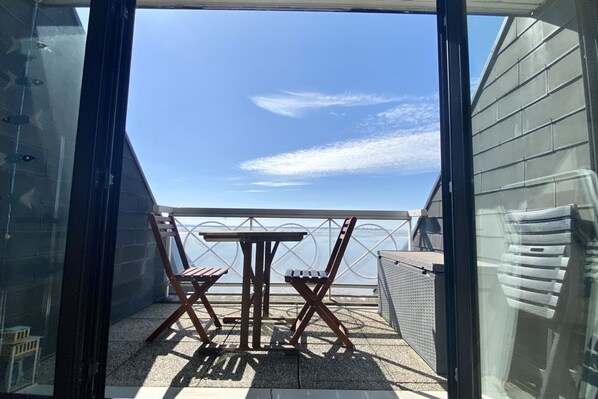 A balcony with a table and two chairs, a view of the sky, and a greenhouse structure in the background.