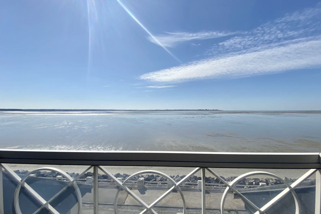 Cielo despejado con el reflejo del sol en el agua, una barandilla metálica y una vista de la costa distante.