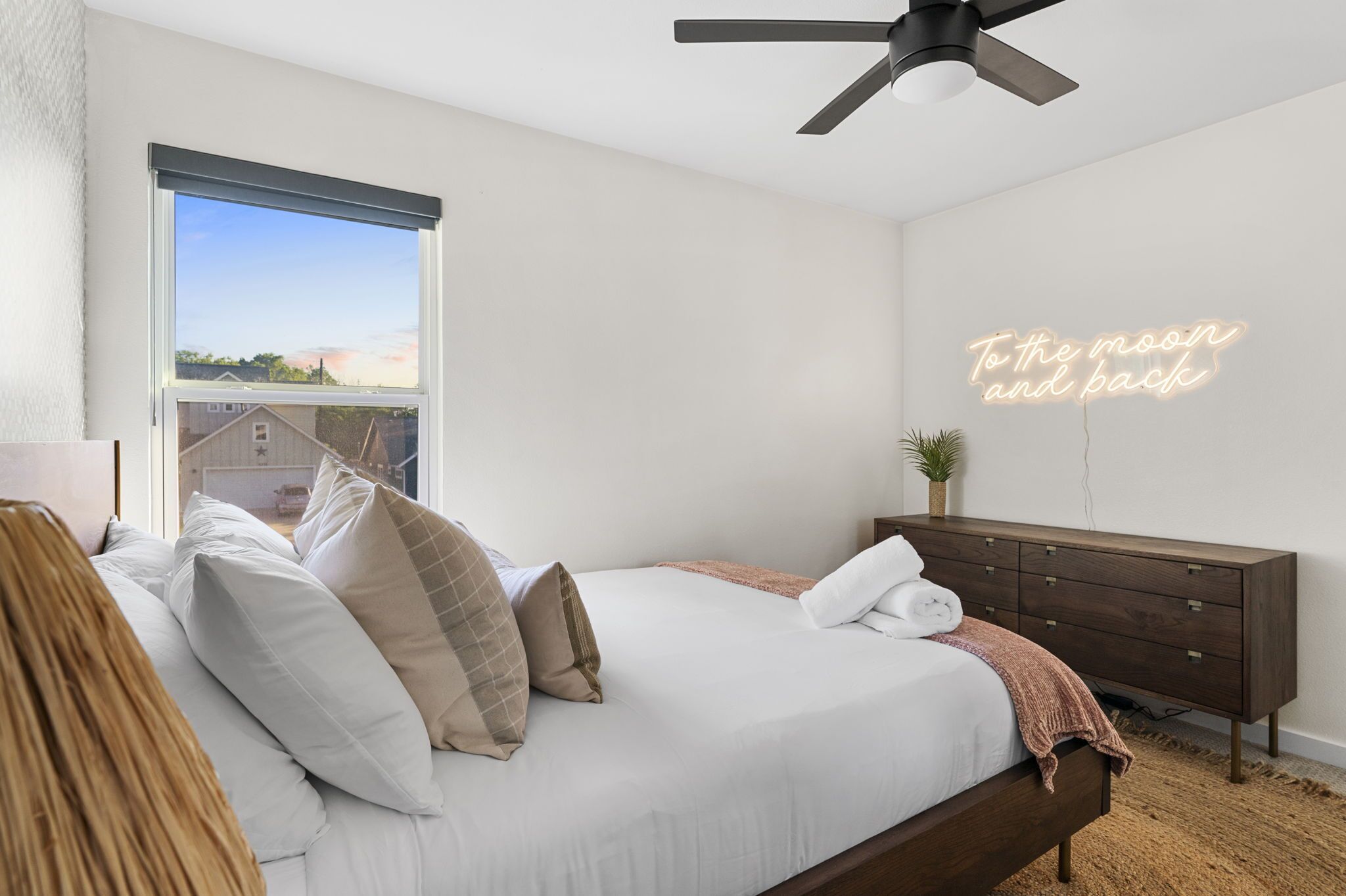 A bedroom with a bed, a ceiling fan, a dresser, a neon sign, and a view of the outside.