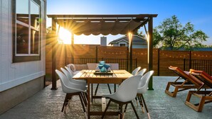 Una terrazza esterna con un tavolo di legno e sedie bianche, una pergola che offre ombra e un recinto di legno sullo sfondo.