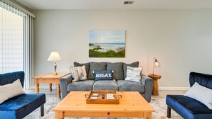 En stue med en grå sofa, blå lænestole, et træbord og et billede af en strand.