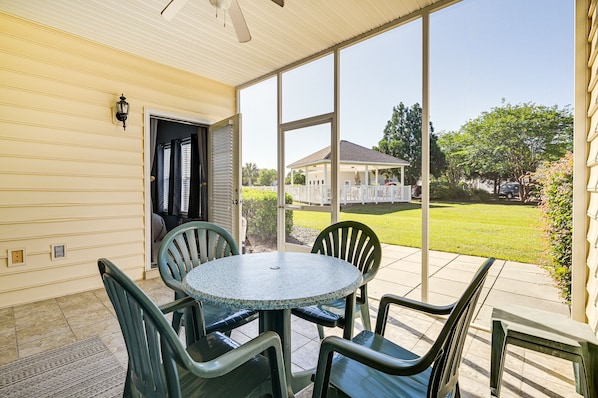 Une terrasse couverte avec une table ronde et des chaises, de grandes fenêtres et une vue sur une maison et une pelouse.