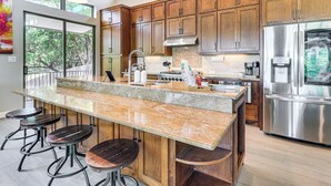 Une cuisine moderne avec des meubles en bois, un grand îlot central doté de tabourets, des appareils électroménagers en acier inoxydable et une vue par la fenêtre.