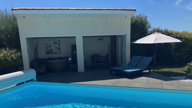 A swimming pool with clear water, a covered patio area with seating, and a white building with an open door. - Gite "bambou" Avec Piscine (Beauvallon)