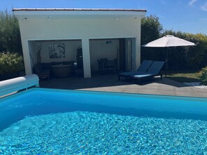 A swimming pool with clear water, a covered patio area with seating, and a white building with an open door. - Gite "bambou" Avec Piscine (Beauvallon)