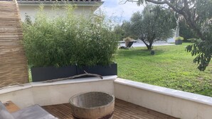 A wooden deck with a cushioned seating area, a rustic barrel, and a view of a well-maintained lawn and trees. - Gite "bambou" Avec Piscine (Beauvallon)