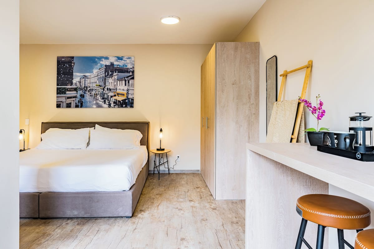 Ein modernes Hotelzimmer mit einem großen Bett, einem kleinen Tisch mit Lampe, einer Bar mit Hockern und einem Stadtbild an der Wand.