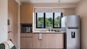 Une cuisine moderne avec des meubles en bois, un réfrigérateur en acier inoxydable, une fenêtre donnant sur un espace verdoyant et un plan de travail doté d’un évier et d’une bouilloire.