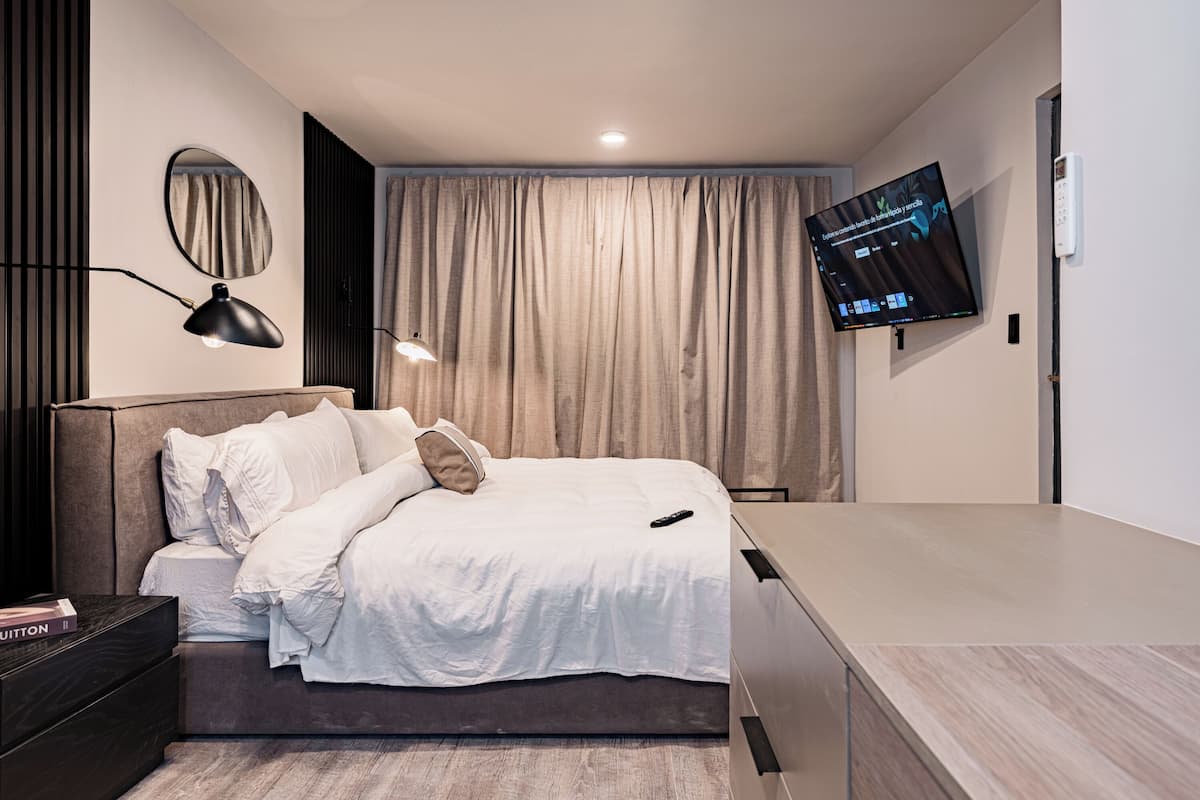 Ein modernes Schlafzimmer mit Bett, Nachttischen, einem an der Wand befestigten Fernseher und einem Fenster mit Vorhängen.