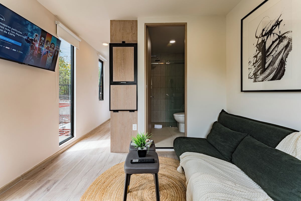 Ein modernes Wohnzimmer mit einem dunklen Sofa, einem kleinen Tisch mit einer Fernbedienung, einer Topfpflanze und einem großen Fenster.
