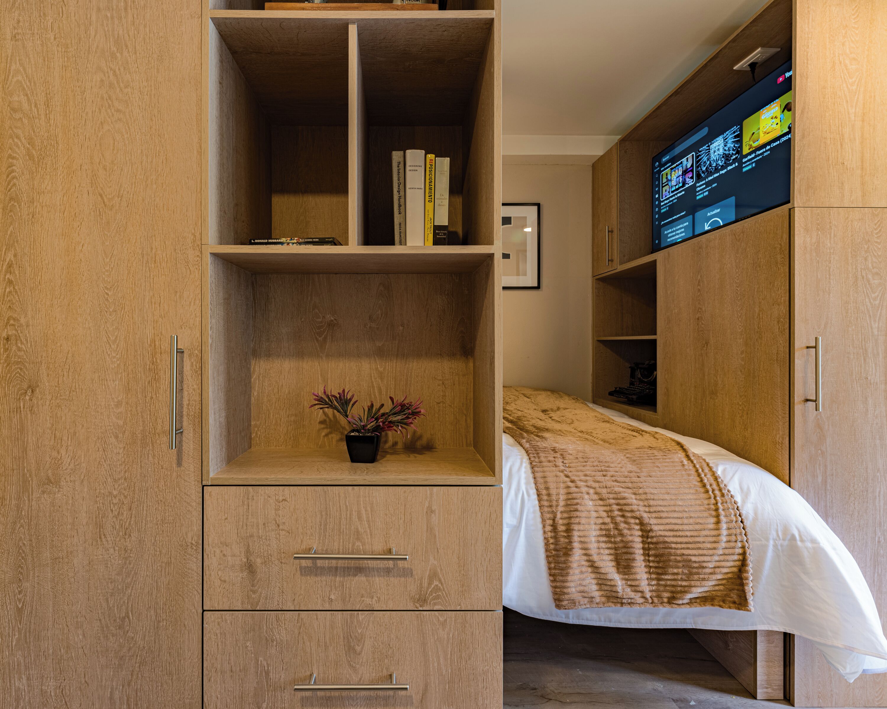 Un dormitorio con un armario de madera, una cama con una manta estampada, una pequeña planta en maceta y un televisor de pared mostrando una interfaz colorida.