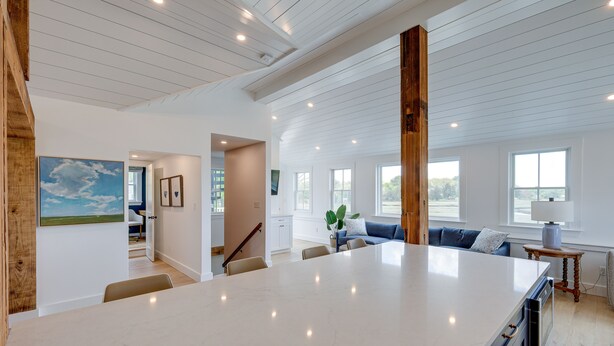 A spacious kitchen with a large island, wooden beams on the ceiling, and a view of the living area with a sofa and windows. - Unique Scituate Vacation Rental on Herring River! (Scituate)