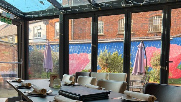 A dining area with a long table set for multiple people, large windows with a view of a brick building and a mural, and a fire exit sign visible. - Oakham hotel (Oakham)