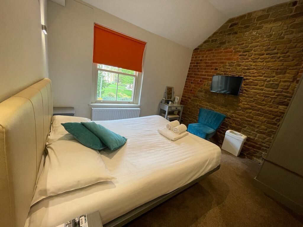 Un dormitorio con cama, ventana con persiana naranja, pared de piedra, televisor montado en la pared y una silla.
