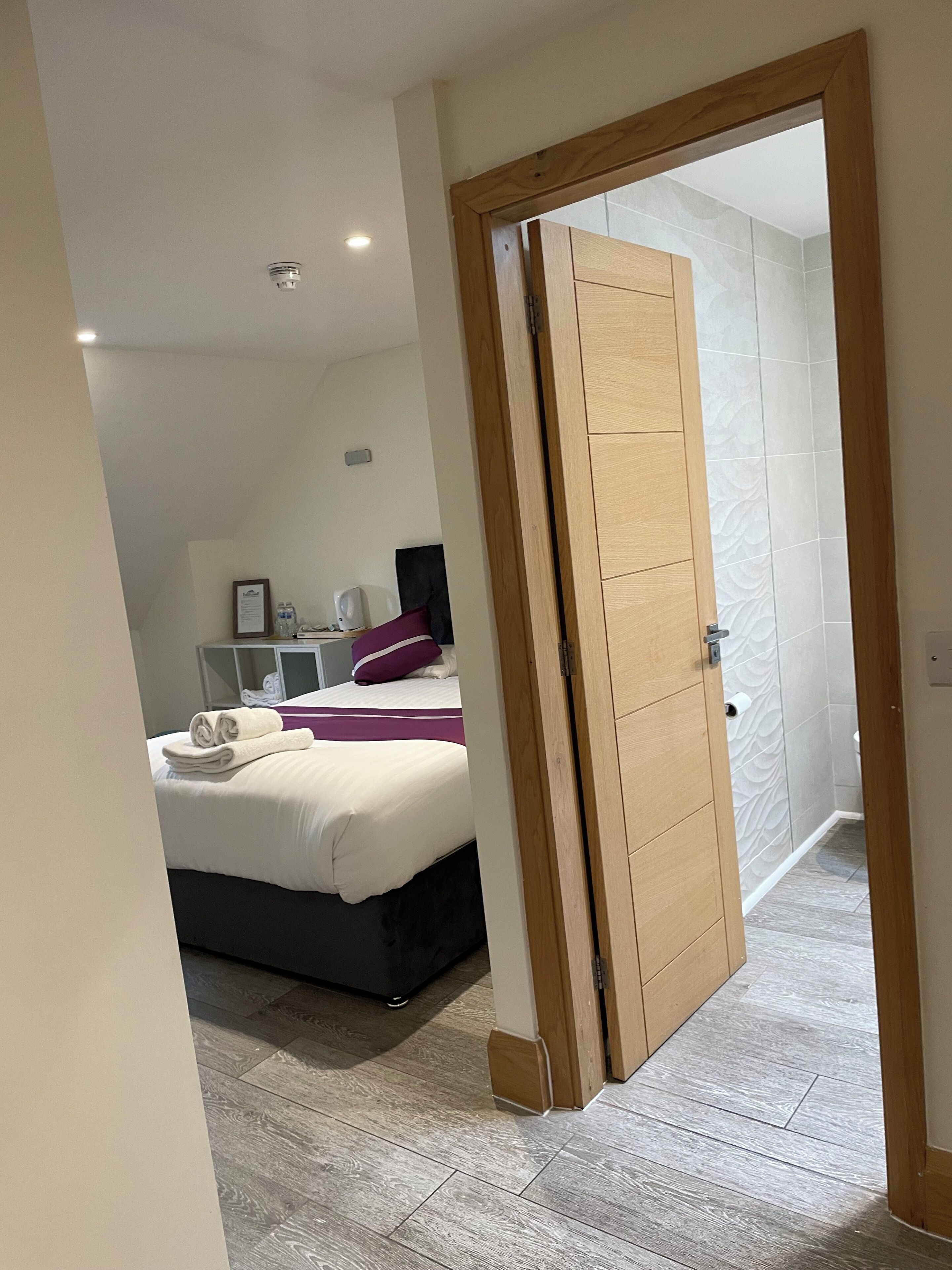 Ein Hotelzimmer mit Bett, Nachttisch, Spiegel und einem durchgehenden Türrahmen sichtbarem Badezimmer.