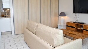 Una sala de estar moderna con un sofá color beige, una mesa de centro de madera, un televisor montado en la pared y una cocina visible a través de una puerta corredera.