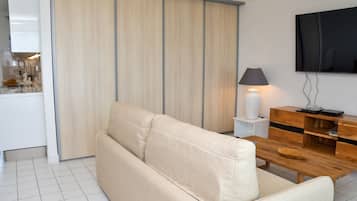 A modern living room with a beige sofa, a wooden coffee table, a TV mounted on the wall, and a kitchen area visible through a sliding door.