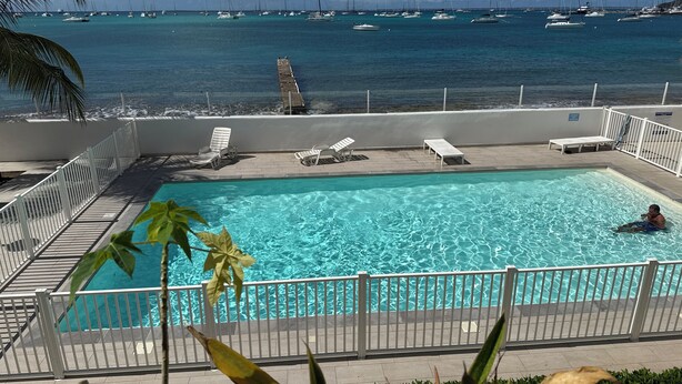 Una piscina con agua cristalina, rodeada de barandillas blancas y sillas de descanso, con vistas a un puerto deportivo lleno de veleros.