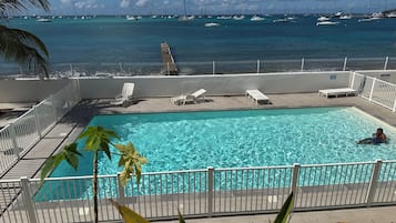 A pool with clear water, surrounded by white railings and lounge chairs, overlooking a marina with sailboats.