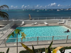 Una piscina con agua cristalina, rodeada de barandillas blancas y sillas de descanso, con vistas a un puerto deportivo lleno de veleros.