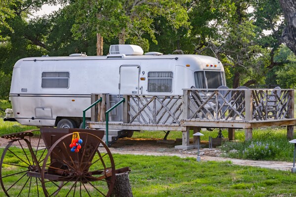 A white RV parked on a grassy area with a wooden deck and a wheelbarrow in the foreground. - The Avion ~ Glamping in Spicewood TX Hill Country at Green Acres (Spicewood)