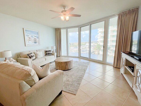 A spacious living room with a large window, a ceiling fan, a sofa set, a coffee table, and a TV. - DOWN BY THE BAY! 3RD FLOOR 2 BEDROOM 2 BATH UNIT!! ASK ABOUT DISCOUNTS! (Orange Beach)