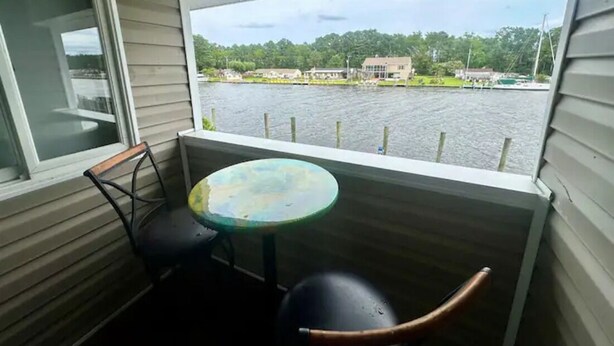 A balcony with a table and chairs, overlooking a body of water with houses and trees in the background. - Knot Two- Balcony View of ICW- Dog Friendly (Coinjock)