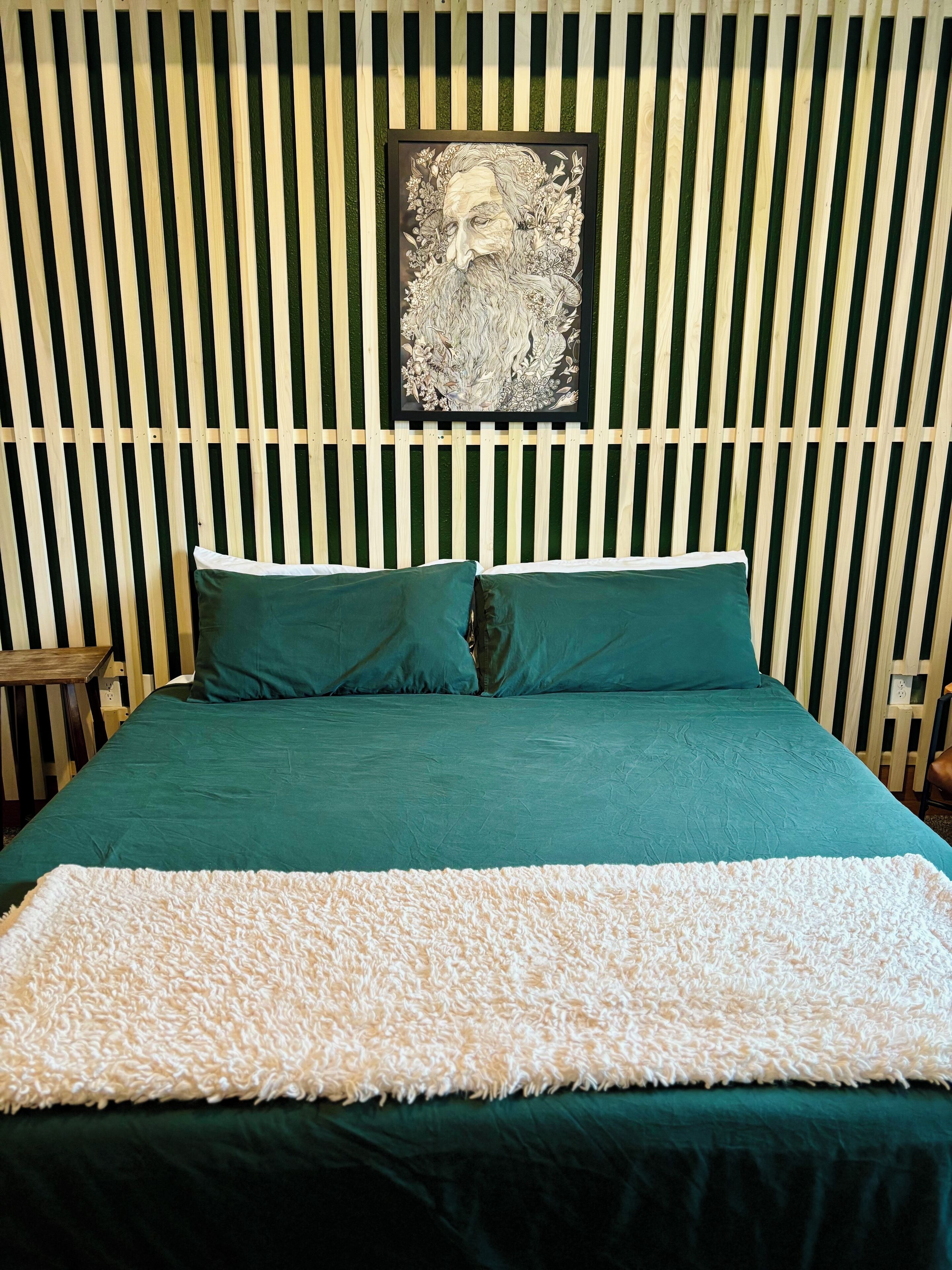 A bed with a green bedspread and two green pillows, a white fluffy throw, and a framed artwork on the wall above.
