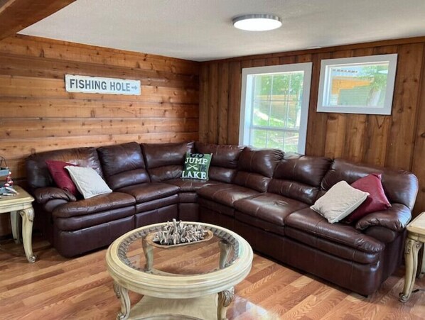A cozy living room with a brown leather sectional sofa, a round coffee table, and a sign that reads "FISHING HOLE". - Tree House Cabin (Avinger)