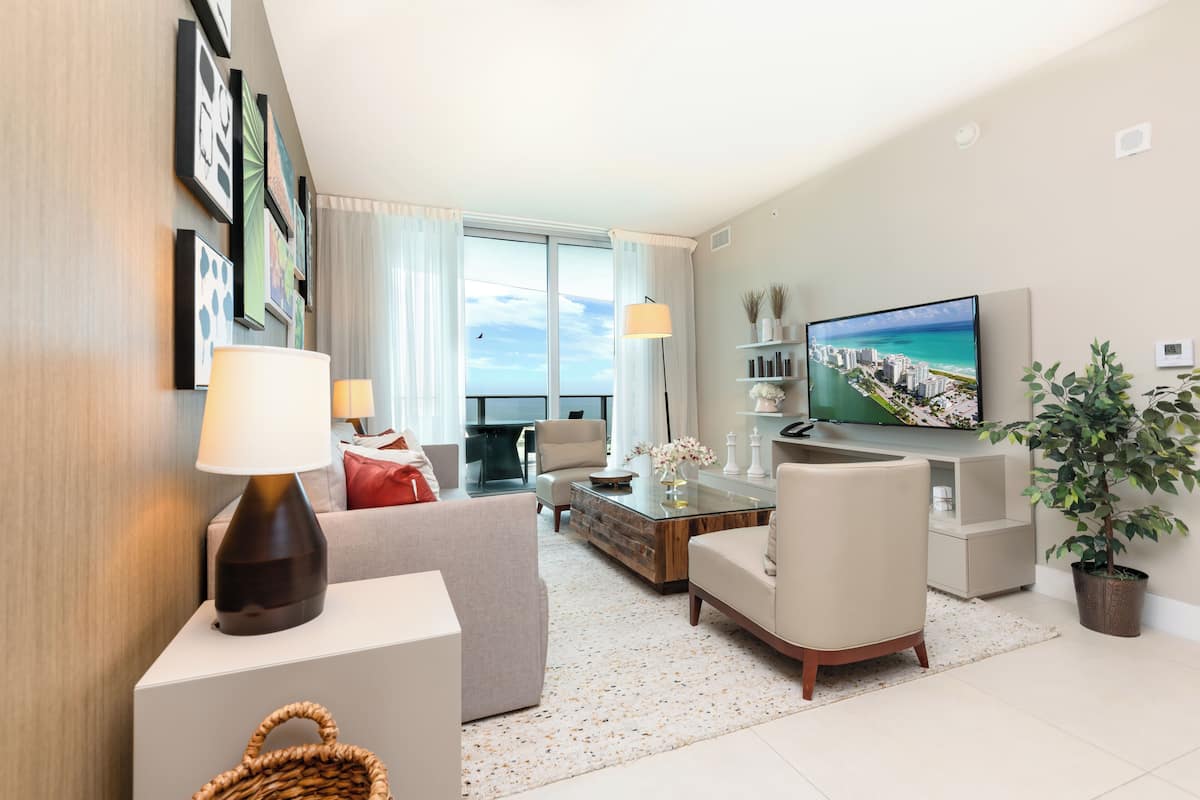 Een moderne woonkamer met een bank, fauteuils, een salontafel en een televisie. Er is een balkon met uitzicht op de oceaan.