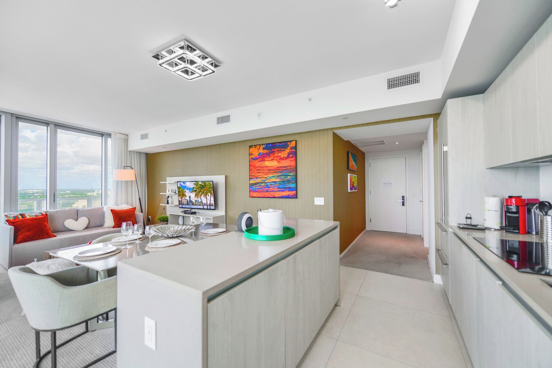 A modern hotel room with a large window, a dining area, a kitchen, and a living area with a TV and artwork.