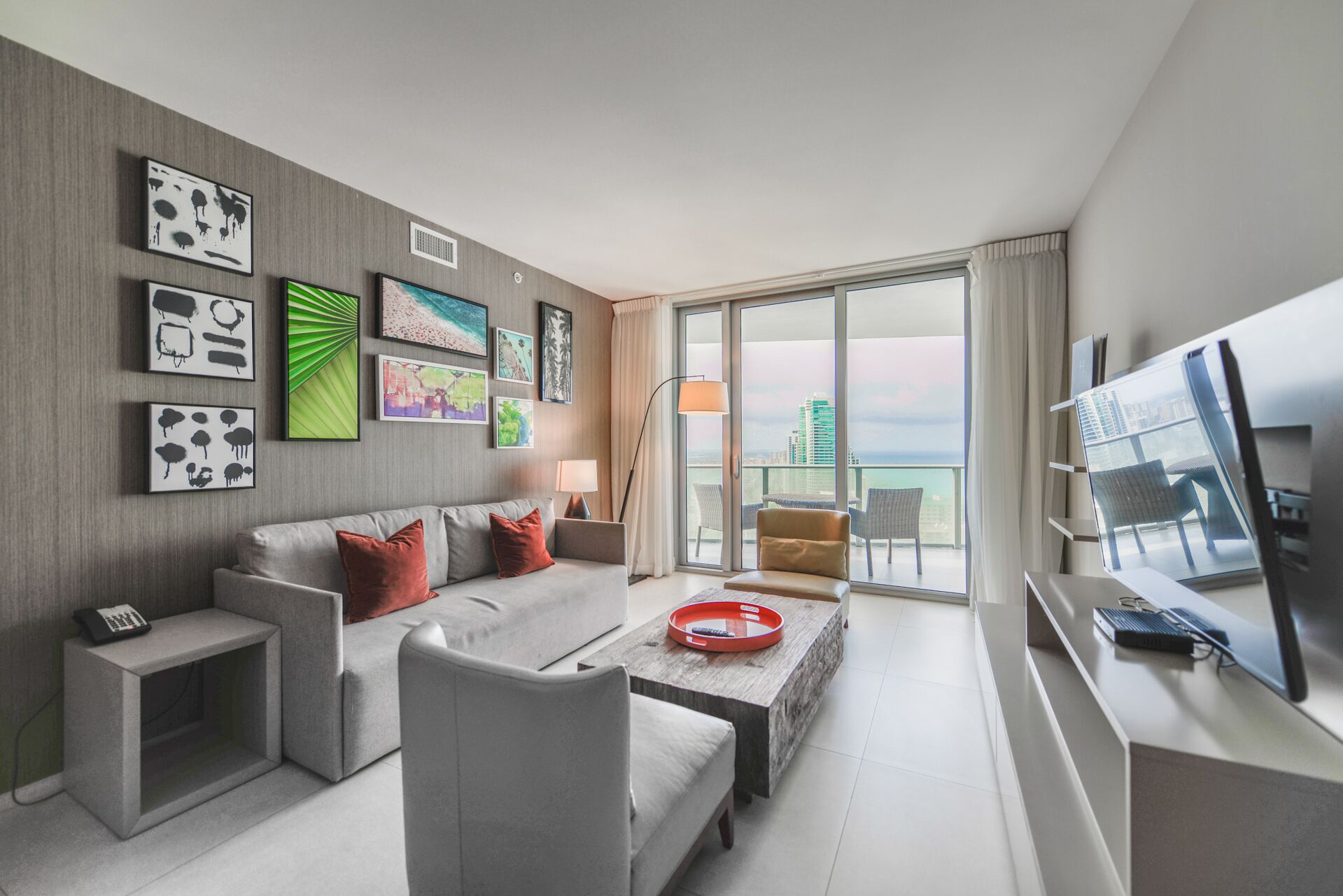 Une chambre d’hôtel moderne avec un canapé gris, une table basse, un téléviseur et des œuvres d’art accrochées aux murs.