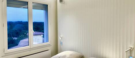 Een slaapkamer met een bed, twee kussens, een raam met uitzicht op groen, en een witte muur met een lichtschakelaar.