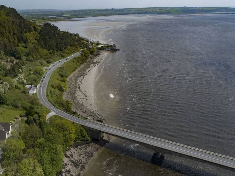 Une route côtière avec un pont enjambant une étendue d’eau, bordée de verdure et de maisons.