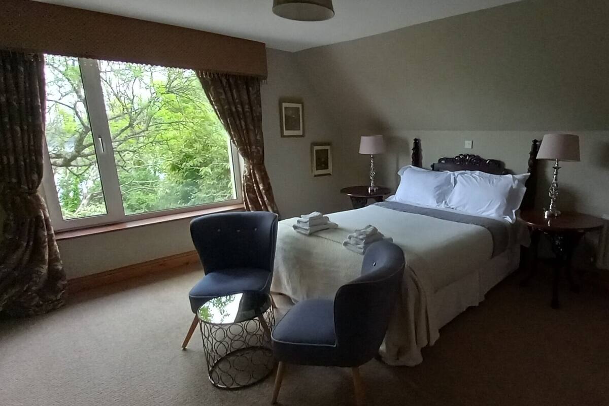 A hotel room with a bed, two chairs, a small table, two bedside lamps, and a window with curtains.