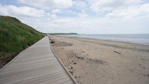 A wooden boardwalk leading to a sandy beach with a grassy hill on one side and a calm sea in the background. - Ardsallagh Lodge (Tinnabinna)