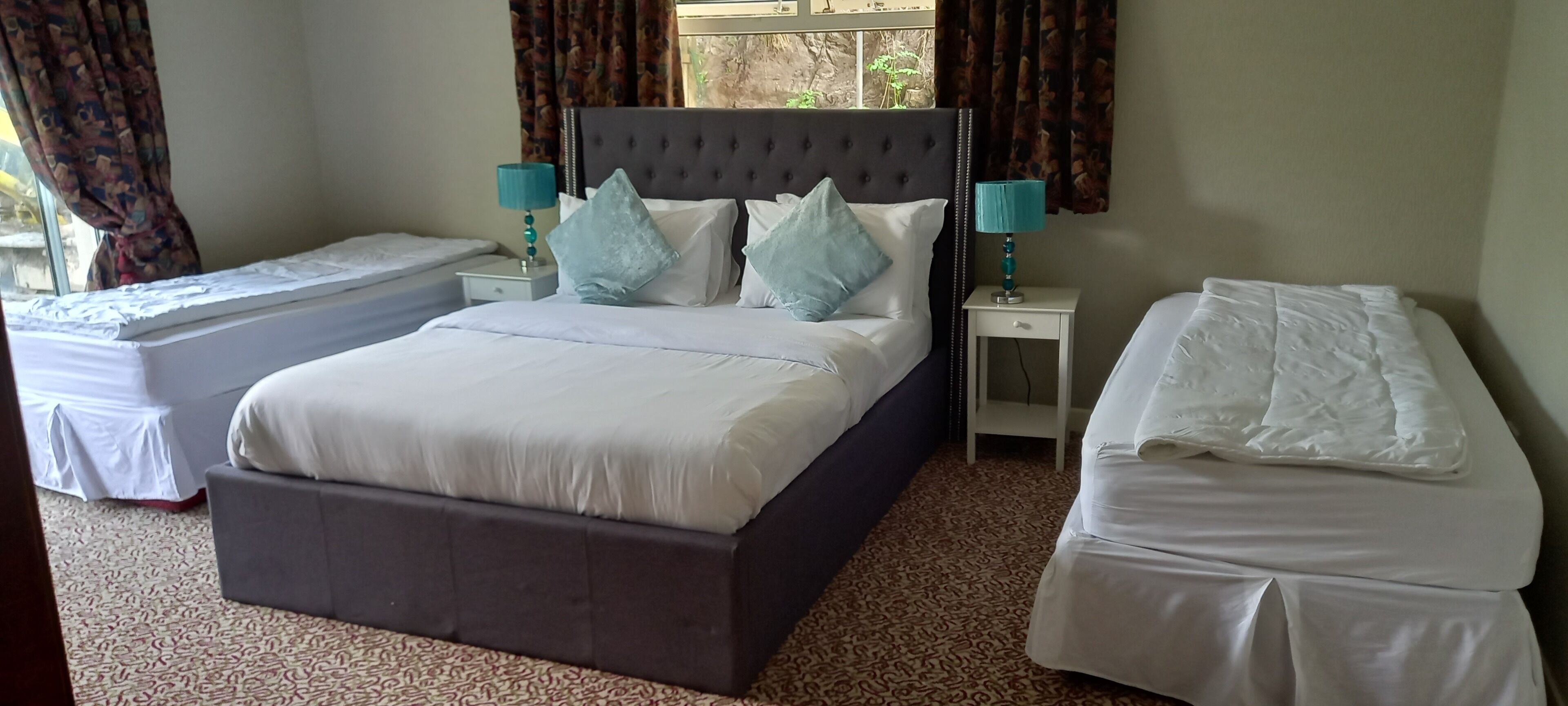 Una habitación de hotel con una cama grande, una cama más pequeña, dos mesitas de noche con lámparas y una ventana con cortinas.