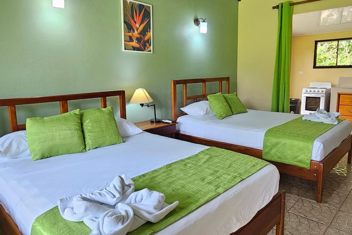 Two double beds with green bedding and white pillows, a nightstand with a lamp, a painting on the wall, and a kitchen area with a stove and green curtains.