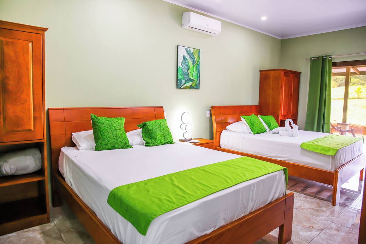 A hotel room with two beds, green pillows, a wooden headboard, a wall-mounted air conditioner, and a window with curtains.